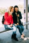 Junge multiethnische Frauen surfen auf dem Smartphone, während sie gemeinsam auf der Bank einer Bushaltestelle sitzen — Stockfoto