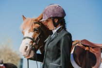 Vista lateral de joven adolescente mujer en casco de jinete y chaqueta acariciando caballo de pie juntos al aire libre contra el cielo azul - foto de stock