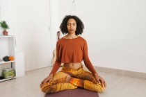 Mujer joven atractiva afroamericana sentada en postura de yoga con las piernas cruzadas y meditando con los ojos cerrados en casa - foto de stock