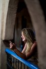 Весела красива жінка в стильному одязі посміхається і переглядає смартфон, спираючись на балконні поручні стародавнього будинку в Марокко. — стокове фото