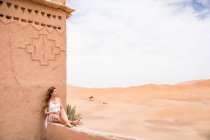 Beautiful young woman in white top sitting on stone fence in wind looking away against endless sandy desert, Morocco — Stock Photo