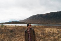 Дорослий чоловік з рюкзаком стоїть у мальовничій віддаленій долині з горами та озером, дивлячись далеко — стокове фото