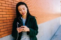 Mujer asiática joven escuchando música y navegando por teléfono inteligente mientras se apoya en la pared de ladrillo en la calle de la ciudad - foto de stock
