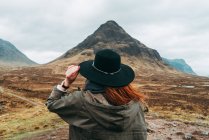 Mujer usando y sosteniendo sombrero de pie contra pintorescas montañas de Escocia - foto de stock