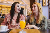 Jóvenes mujeres multirraciales sonriendo y hablando entre sí mientras se sientan en la mesa en el acogedor café - foto de stock