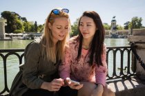 Молодые многорасовые женщины в модных нарядах улыбаются и просматривают смартфон, сидя рядом с набережной в солнечный день — стоковое фото