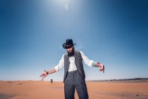 Hombre barbudo disfrazado de vaquero mirando hacia abajo mientras está de pie en el desierto contra el cielo azul - foto de stock