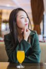 Cher asiatique femme en élégant manteau vert regardant loin tout en étant assis à la table de café avec un verre de jus frais — Photo de stock