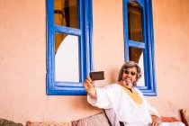 Веселий дорослий чоловік у довгому одязі сидить на дивані й користується телефоном у домі, прикрашеному східним стилем (Марокко). — стокове фото