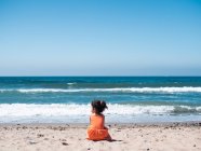 Back view of female kid in orange dress sitting on sandy seaside and looking at sea and sky — Stock Photo
