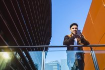 Hombre positivo en traje elegante usando el teléfono móvil mientras está de pie en el balcón de cristal moderno del edificio contemporáneo en el día soleado - foto de stock