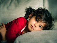 Adorable enfant féminin en robe rouge souriant tout en se reposant sur l'oreiller à la maison — Photo de stock