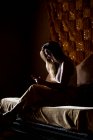 Side view of young lady smiling and using smartphone while sitting on soft bed in dark room in Morocco — Stock Photo