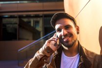 Позитивний чоловік у стильному вбранні розмовляє на мобільному телефоні, спираючись на стіну сучасної будівлі в сонячний день — стокове фото