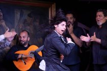 Hombre en traje negro bailando flamenco cerca de músicos masculinos hispanos durante la actuación contra la pintura en el escenario oscuro - foto de stock
