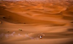 Entfernte Luftaufnahme von fröhlich stilvolle Brünette mit erhobenem Arm mit brennenden rauchigen Feuerwerk steht in der Wüste von Marokko — Stockfoto