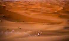 Distant aerial view of cheerful stylish brunette holding arm raised with burning smoky firework standing in desert of Morocco — Stock Photo