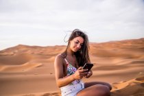 Mujer joven usando el teléfono mientras está sentada en medio del desierto de arena, Marruecos - foto de stock