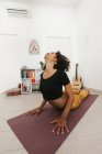 Mujer joven afroamericana en postura de yoga con la cabeza hacia abajo estirándose en la estera en la sala de luz - foto de stock