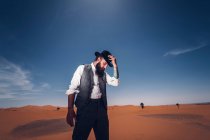 Homem barbudo em traje de cowboy olhando para baixo enquanto estava no deserto contra o céu azul — Fotografia de Stock