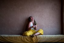 Mujer rubia feliz sentada en el sofá en la terraza en estilo oriental y usando un teléfono móvil en Marruecos - foto de stock