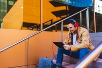 Bello uomo in cuffia ascoltare musica e tablet di navigazione mentre seduto sulle scale fuori edificio moderno — Foto stock