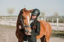 Вид сбоку на молодую девушку-подростка в шлеме жокея и ласкающую лошадь, стоящую на улице — стоковое фото