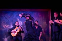 Woman in black costume dancing flamenco near Hispanic male musicians during performance against painting on dark stage — Stock Photo