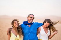Hombre barbudo mayor riendo y abrazando a mujeres alegres mientras camina en el desierto de arena durante su viaje a Marruecos - foto de stock