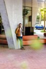 Confiante jovem na moda inclinando-se sobre pilar de concreto perto de sofá de couro fora edifício contemporâneo na rua da cidade — Fotografia de Stock