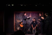 Homem de traje preto dançando flamenco perto de músicos masculinos hispânicos durante a performance contra a pintura no palco escuro — Fotografia de Stock