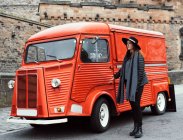 Redhead woman passing though a retro red colored van on roadside with aged stone castle on background, Scotland — Stock Photo