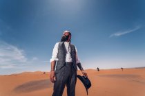Homem barbudo em traje de cowboy olhando para longe enquanto estava no deserto contra o céu azul — Fotografia de Stock