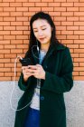Mujer asiática joven escuchando música y navegando por teléfono inteligente mientras se apoya en la pared de ladrillo en la calle de la ciudad - foto de stock