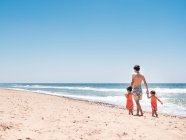 Vista posteriore dell'uomo che cammina lungo il mare con le bambine sullo sfondo del mare calmo e del cielo blu — Foto stock