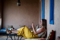 Mujer adulta sentada en un sofá en una terraza de estilo oriental y usando un teléfono móvil en Marruecos - foto de stock