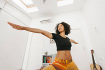 Mujer joven afroamericana realizando postura de yoga con brazos extendidos en sala de luz - foto de stock