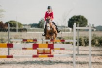 Jockey adolescente a caballo saltando sobre barras horizontales de madera mientras monta en pista de carreras - foto de stock