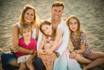 Adulto homem amoroso e mulher com filho e filhas sentados juntos na praia na parte traseira iluminada sorrindo para a câmera — Fotografia de Stock