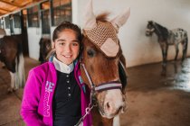 Bambina abbracciata con piccolo pony in cappello carino sulle orecchie in piedi all'interno della stalla e guardando la fotocamera — Foto stock