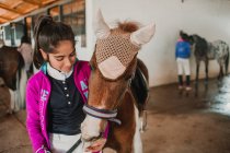 Bambina che abbraccia con piccolo pony in cappello carino sulle orecchie in piedi all'interno della stalla — Foto stock