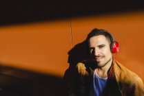 Positive young man in stylish outfit with headphones listening to music near wall with shadow — Stock Photo
