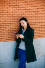 Молодая азиатка слушает музыку и просматривает смартфон, опираясь на кирпичную стену на городской улице — стоковое фото
