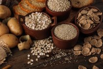 De arriba cereales semillas de avena en cuencos patatas y pan en rodajas sobre fondo de madera - foto de stock