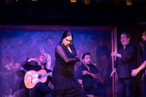 Mulher de traje preto dançando flamenco perto de músicos masculinos hispânicos durante a performance contra a pintura no palco escuro — Fotografia de Stock