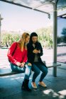 Молоді багаторасові жінки переглядають смартфон, сидячи на лавці автобусної зупинки разом — стокове фото