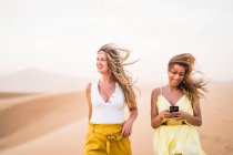 Allegro elegante due amiche bionde che utilizzano il telefono cellulare mentre camminano nel deserto del Marocco — Foto stock