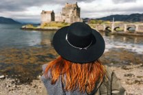 Vista trasera de una mujer elegante con un sombrero contemplando el viejo castillo de piedra en la costa en las montañas, Escocia - foto de stock