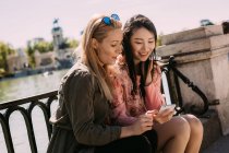 Молоді багаторасові жінки в модних вбраннях посміхаються і переглядають смартфон, сидячи біля набережних перил в сонячний день на вулиці міста — стокове фото