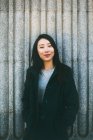 Юна азіатка в елегантному пальто посміхається і дивиться на камеру, спираючись на мармурову стіну на вулиці міста — стокове фото